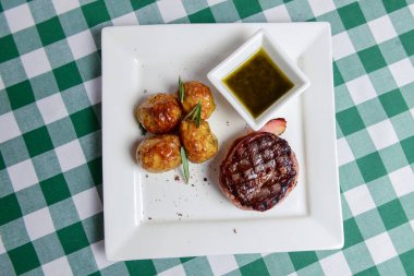 Izgara biftek, fırında patates ve sosla servis edilmiş yeşil ekose masa örtüsünün üzerinde beyaz bir tabak. Barbekü, barbekü konsepti. Lezzetli yaz yemeği.