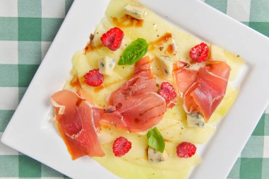 Lezzetli aperatif, peynir ve jambonlu salata. Geleneksel İtalyan mutfağı, İtalyan konsepti. Kare beyaz bir tabakta servis edilen yeşil ekose masa örtüsü..