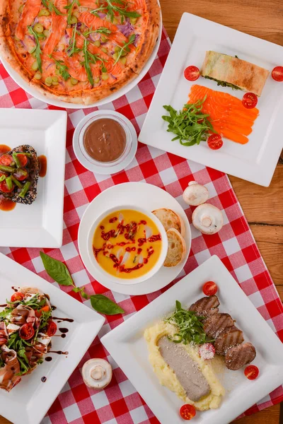 Flat lay shot of delicious italian cuisine. Delicious pizza, salmon rolls, pumpkin soup and salad on a wooden table with plaid tablecloth. — Stock Image Flat Lay Shot Delicious Italian Cuisine Delicious Pizza Salmon Rolls Stock Image