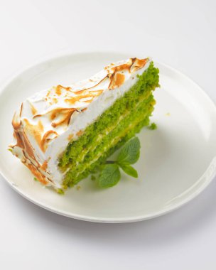 Bir parça yeşil çay keki. Beyaz arka planda beyaz tabağın etrafında servis edilen krema kremalı ve nane yaprağıyla bir parça kibrit çayı keki. Çay ya da kahve için lezzetli bir tatlı..