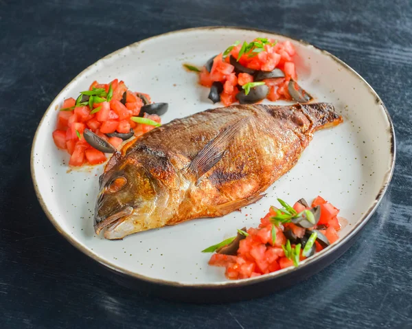 新鮮なハーブ トマト オリーブで揚げた魚は 黒素朴な木製のテーブルの背景に白いプレートで提供しています ディナーの食事 伝統的な食品 お皿の上の焼き魚 — ストック写真