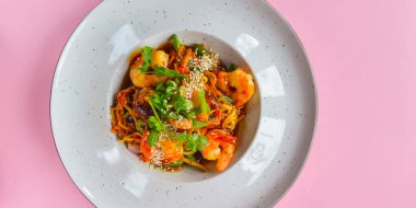 Karides, kuşkonmaz ve susamlı Asya eriştesi. Lezzetli karides, geleneksel Asya mutfağı. Pastel pembe arka planda beyaz bir tabakta servis edilmiş. Boşluk afişini kopyala.