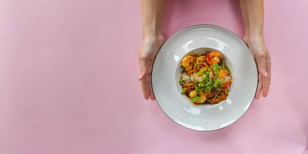 Asian noodles with shrimps, asparagus and sesame seeds. Delicious shrimp wog, traditional Asian cuisine. Served in a white plate over pastel pink background. Copy space banner.