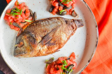 Siyah rustik ahşap masa zemininde beyaz bir tabakta taze otlu, domatesli ve zeytinli kızarmış balık servis ediliyor. Akşam yemeği, geleneksel yemekler. Tabakta tam kızarmış balık..