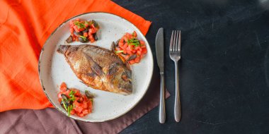 Siyah rustik ahşap masa zemininde beyaz bir tabakta taze otlu, domatesli ve zeytinli kızarmış balık servis ediliyor. Akşam yemeği, geleneksel yemekler. Tabakta tam kızarmış balık..