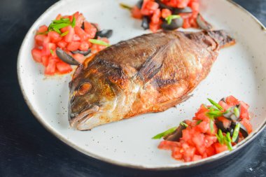 Siyah rustik ahşap masa zemininde beyaz bir tabakta taze otlu, domatesli ve zeytinli kızarmış balık servis ediliyor. Akşam yemeği, geleneksel yemekler. Tabakta tam kızarmış balık..