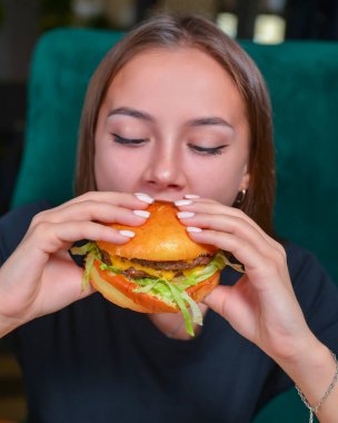 Hamburger yiyen ve patates kızartması yiyen bir kadın. Kafkasyalı güzel bir kadın model elleri bulanık bir restoranın arka planında hamburger yiyor. Hayat, dışarıda yemek kavramı..