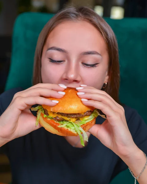 Woman eating burger and fries smiling. Beautiful caucasian female model eating a hamburger with hands over blurred restaurant background. Still life, eating out concept.