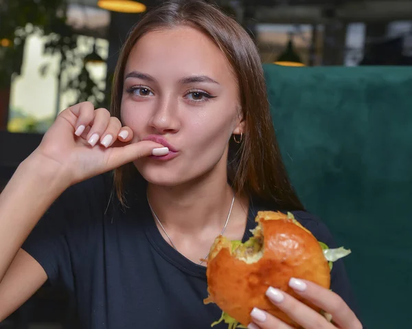 Woman eating burger and fries smiling. Beautiful caucasian female model eating a hamburger with hands over blurred restaurant background. Still life, eating out concept.