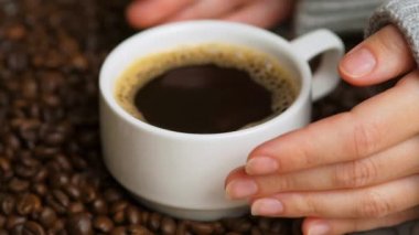 Kahve çekirdeği arka planında bir fincan koyu kahve olan ellerin yan görüntüsü. Bir fincan sade kahve. Nazik kadın elleri küçük bir fincan kahve alıyor. Seçici odak, kapat.