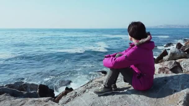 Jeune femme caucasienne avec manteau rose assis sur les rochers près de la mer ondulée — Video