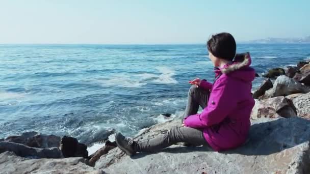 Giovane donna caucasica con cappotto rosa seduta sulle rocce vicino al mare ondulato — Video Stock