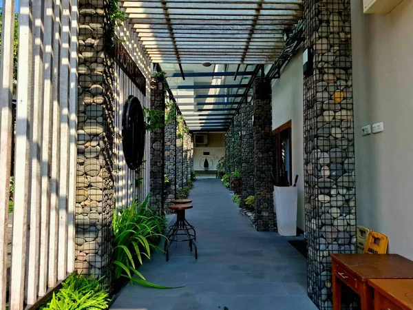 Otel misafirleri için bir koridor, doğal taş sütunları ve sanatsal ahşap pergola çatısı olan. Aydınlık bir sabahta. Göz düzeyi görünümü.