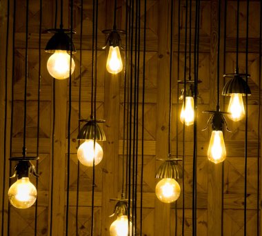 Çoklu vintage tarzı asma ampulleri ahşap panelli bir duvarı aydınlatır. Açığa çıkan ampullerin sıcak kehribar parıltısı, kafeler, barlar veya endüstriyel ve retro estetik içerikli iç tasarım temaları için ideal, rahat ve rustik bir atmosfer yaratır..