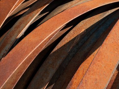 Üst üste binen paslı metal şeritlerin detaylı bir yakın çekimi, güneş ışığıyla aydınlatılan zengin kahverengi ve turuncu renkte yıpranmış yüzeyleri gösteriyor. Soyut düzenleme pas dokusunu, renk değişimlerini ve organik desenleri vurgular.