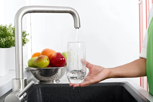 The girl fills a glass with water from a water tap in the kitchen.