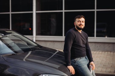 Genç adam arabanın kaputunda oturmuş içkisini içiyor.