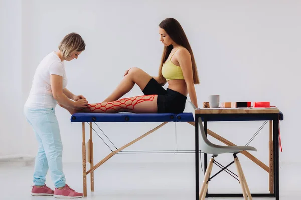 Female doctor helps woman by putting kinesio tape on her leg. Young caucasian girl with kinesiology elastic therapeutic tape on her leg