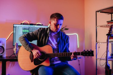 Evde akustik gitar çalan, bilgisayar, mikrofon ve ses ekipmanları kullanan genç bir müzisyen.