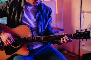 Akustik gitarını renkli neon ışıklarla aydınlatılmış bir stüdyoda çalan genç bir müzisyen.