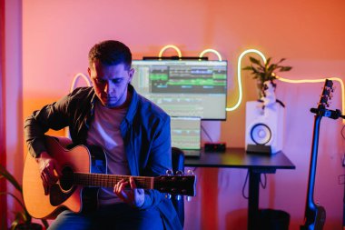 Genç adam akustik gitar çalıyor ve evde bilgisayar ve müzik prodüksiyon yazılımı kullanarak yeni bir şarkı besteliyor.