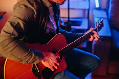 Genç adam renkli ışıkları olan rahat bir ev ortamında akustik gitar çalıyor.