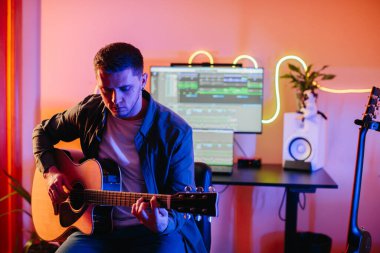 Yeni müzik besteleyen genç müzisyen evdeki stüdyoda bilgisayar ve neon ışıklarla akustik gitar çalıyor.