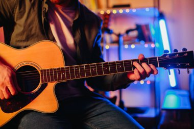 Renkli ışıklarla aydınlatılmış rahat bir ev stüdyosunda akustik bir gitarla müzik besteleyen bir müzisyenin yakın çekimi.