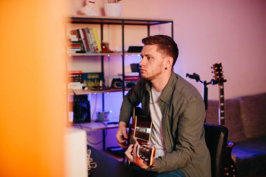 Genç müzisyen yaratıcı ilham kaynağıyla çevrili rahat bir ev stüdyosunda akustik gitar çalarken orijinal şarkılar besteledi.