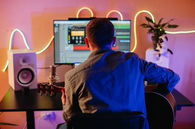 Gitar çalarken melodi besteleyen ve modern ekipmanlarla dolu rahat bir ev stüdyosunda bilgisayar yazılımı kullanan genç bir müzisyen.