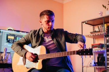 Genç bir adam, yaratıcı enerjiyle dolu rahat bir ev stüdyosunda akustik bir gitarı akort ediyor.