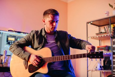 Bir ev stüdyosunda müzik besteleyen genç adam, akustik gitar melodilerini bilgisayarda dijital seslerle ustalıkla harmanlıyor.