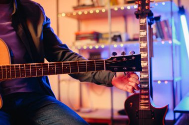 Canlı bir ev kayıt stüdyosunda akustik gitar akort eden bir müzisyenin yakın çekim görüntüsü, renkli ışıklar ve yaratıcı enerjiyle çevrili.