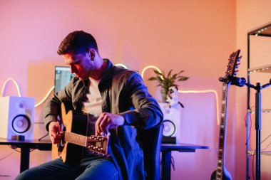 Genç adam akustik gitar çalıyor ve modern ekipman ve canlı renklerle çevrili rahat bir ev kayıt stüdyosunda müzik besteliyor.