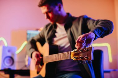 Genç müzisyen akustik gitarını akort ediyor ve renkli neon ışıklarıyla ev stüdyosunda müzik besteliyor.