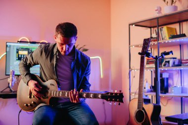 Yaratıcı ekipman ve ilham kaynağıyla çevrili rahat bir ev stüdyosunda elektro gitar çalarken orjinal müzik besteleyen genç bir müzisyen.