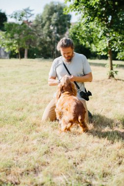Fotoğrafçı köpeğinin tasmasını güneşli bir parkta eğip düzeltiyor ve evcil hayvanıyla huzurlu bir anın tadını çıkarıyor.