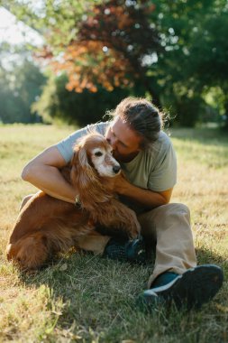 Çimenlerde oturan adam, güneşli bir parkta sevgili cocker köpeğini kucaklıyor ve öpüyor, birlikte neşeli bir anın tadını çıkarıyor.