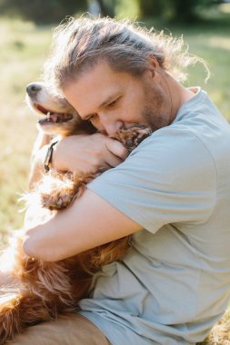 Olgun bir adam, güneşli bir parkta kürklü arkadaşını kucaklıyor. Yumuşak bir bağ ve şefkat anının tadını çıkarıyor.