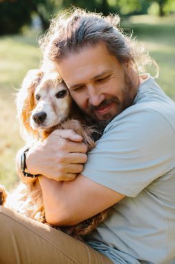 Orta yaşlı bir adam, sakin bir park ortamında tüylü arkadaşını nazikçe kucaklıyor, bir anlık bağlantının ve sevginin tadını çıkarıyor.