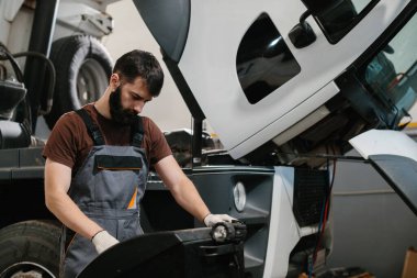 Otomobil tamircisi garajda bir kamyon motoru üzerinde çalışıyor. Aracı incelemek ve onarmak için el feneri ve aletler kullanıyor.