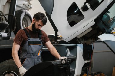 Tamirci iş tulumu giyerek garajdaki bir kamyon motorunu inceler ve en uygun araç performansı için onarım ve bakım üzerine odaklanır.