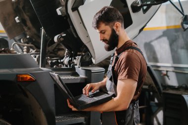 Otomobil tamircisi, garajdaki bir kamyon motorunu teşhis etmek ve onarmak için dizüstü bilgisayar kullanıyor. Araç bakımında modern teknolojiyi gösteriyor.