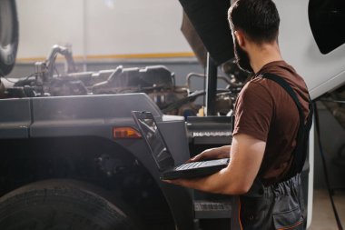 Oto tamircisi, yoğun bir tamirhanede bir kamyon motorunu incelerken, etkin bakım ve servis sağlarken, sistem kontrolü için bir dizüstü bilgisayar kullanıyor.