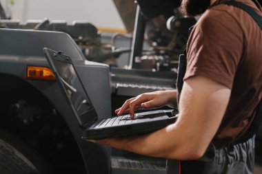 Otomobil tamircisi modern bir tamirhanede dizüstü bilgisayar kullanarak kamyon teşhisi üzerinde çalışıyor, verimli ve kusursuz araç bakımı sağlıyor.