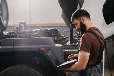 Otomobil tamircisi bir garajda kamyon motoru sorunu teşhis etmek için dizüstü bilgisayar kullanıyor, bakım ve onarım yapıyor.