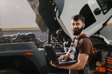 Bir atölyedeki oto tamircisi, bir kamyonu teşhis etmek ve tamir etmek için dizüstü bilgisayar kullanıyor. Arka planda görünen kaput ve atölye araçlarını aç