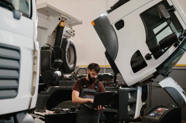 Otomobil tamircisi garajda bir kamyon arızası teşhis ederken dizüstü bilgisayar kullanıyor, araç tamirinde uzmanlık gösteriyor