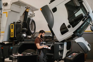 Otomobil tamircisi garajda bir kamyon motoru üzerinde çalışıyor. Onarım işlemi sırasında dizüstü bilgisayar kullanıyor.