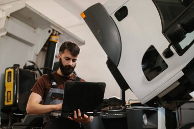 Otomobil tamircisi garajda bir kamyonu incelerken dizüstü bilgisayar kullanıyor.